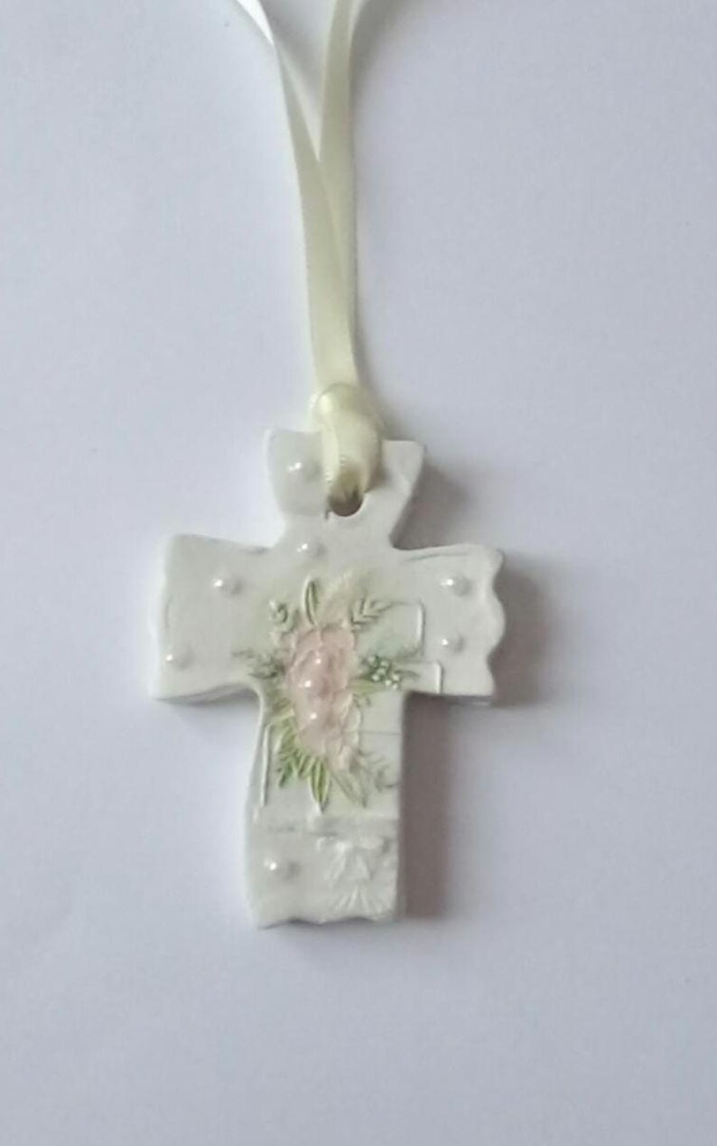 May include: White ceramic cross pendant with a floral design in pink and green. The cross has a ribbon for hanging and is decorated with small, raised dots. A delicate religious ornament.