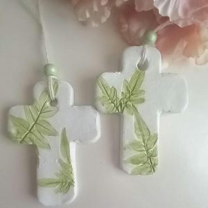 May include: Two white, cross-shaped ornaments with green leaf imprints. Each ornament has a small hole at the top with a string and a green bead for hanging. The background features soft pink and white flowers.