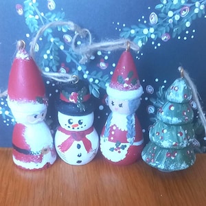 May include: Four hand-painted wooden Christmas ornaments featuring a Santa Claus, a snowman with a black top hat, a figure with braided hair in a red dress, and a green Christmas tree. Each ornament includes a jute twine hanging loop.