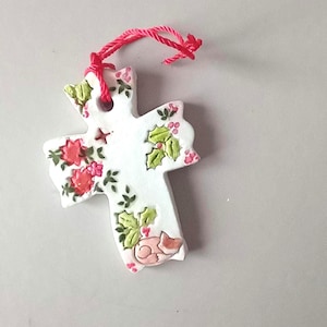 May include: A white ceramic cross ornament featuring hand-painted red flowers, green holly leaves, and a small sleeping fox at the base. The cross has a hole at the top with a bright red string attached for hanging purposes.
