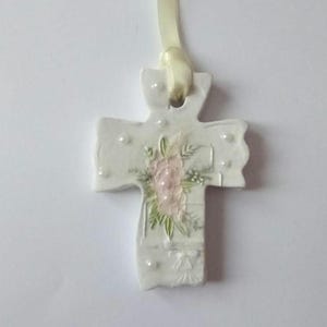 May include: A white ceramic cross pendant with a floral design in pink and green. The cross is adorned with small, pearl-like embellishments and a cream-colored ribbon for hanging. A delicate religious ornament.