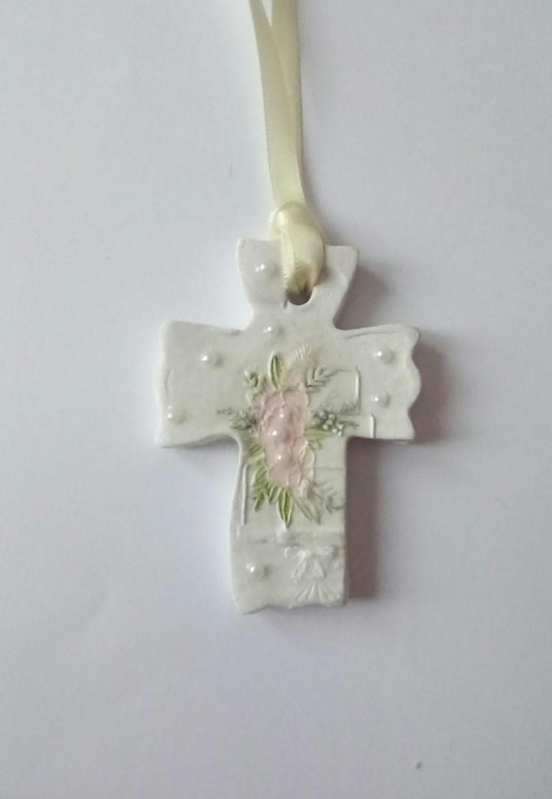 May include: A white ceramic cross pendant with a floral design in pink and green. The cross is adorned with small, pearl-like embellishments and a cream-colored ribbon for hanging. A delicate religious ornament.