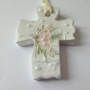 May include: A white cross-shaped ornament with a floral design. The cross features a pink flower arrangement with green leaves and small white pearl accents. A cream-colored ribbon is attached for hanging.