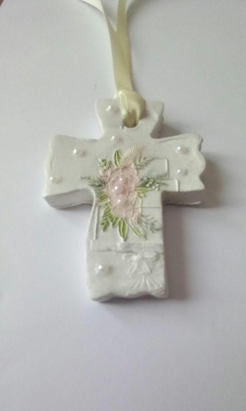 May include: A white cross-shaped ornament with a floral design. The cross features a pink flower arrangement with green leaves and small white pearl accents. A cream-colored ribbon is attached for hanging.