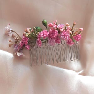 May include: A hair comb featuring a cluster of small pink flowers, buds, and green stems attached to a silver-toned metal base. The arrangement includes various shades of pink blossoms and small, rounded green buds, creating a dense floral display.