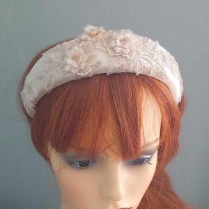 May include: A white satin headband with delicate lace and floral embroidery. The headband is wide and has a curved shape.