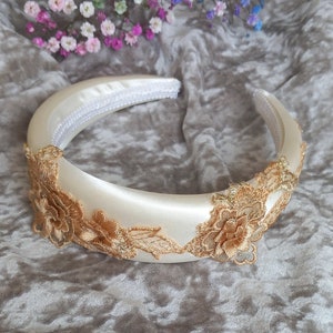 May include: A white satin headband with gold lace detailing. The lace features delicate floral designs.
