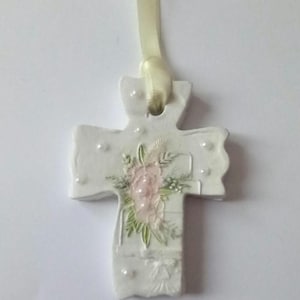 May include: A white ceramic cross pendant with a floral design in pink and green. The cross is adorned with small, raised dots and hangs from a cream-colored ribbon. A religious-themed item.