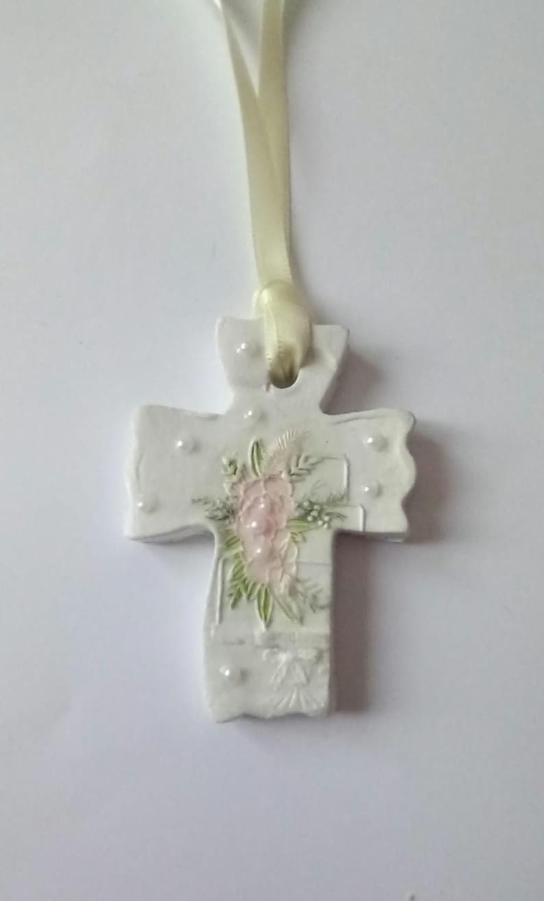 May include: A white ceramic cross pendant with a floral design in pink and green. The cross is adorned with small, raised dots and hangs from a cream-colored ribbon. A religious-themed item.