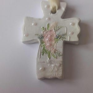 May include: A white cross-shaped ornament with a floral design and pearl accents. The cross features a pink and green floral arrangement and is suspended by a cream-colored ribbon. A decorative item.