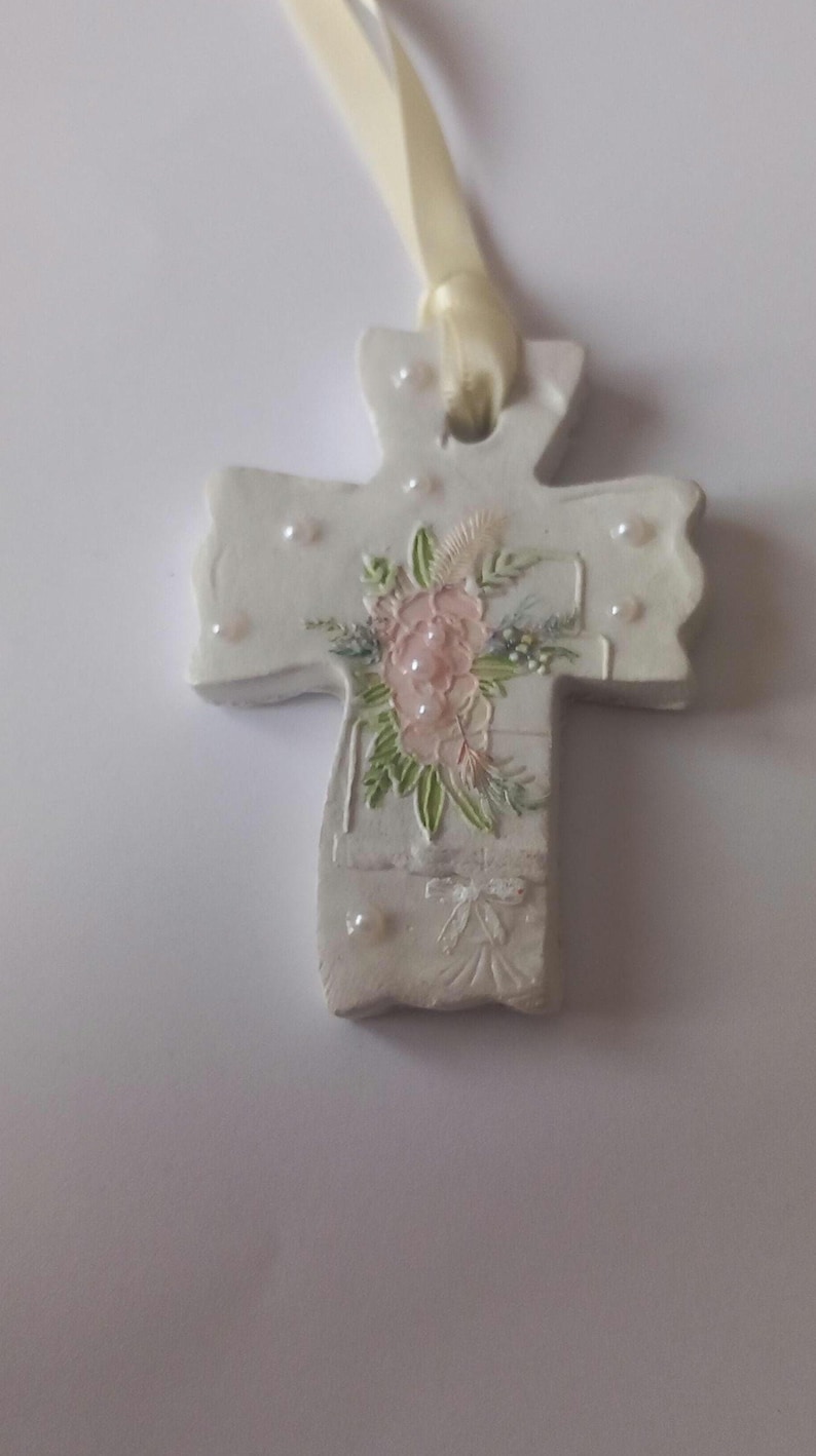 May include: A white cross-shaped ornament with a floral design and pearl accents. The cross features a pink and green floral arrangement and is suspended by a cream-colored ribbon. A decorative item.