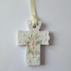 May include: A white ceramic cross pendant with a cream-colored ribbon. The cross is decorated with a pink floral design and small white pearl accents. The pendant is approximately 3 inches in length.