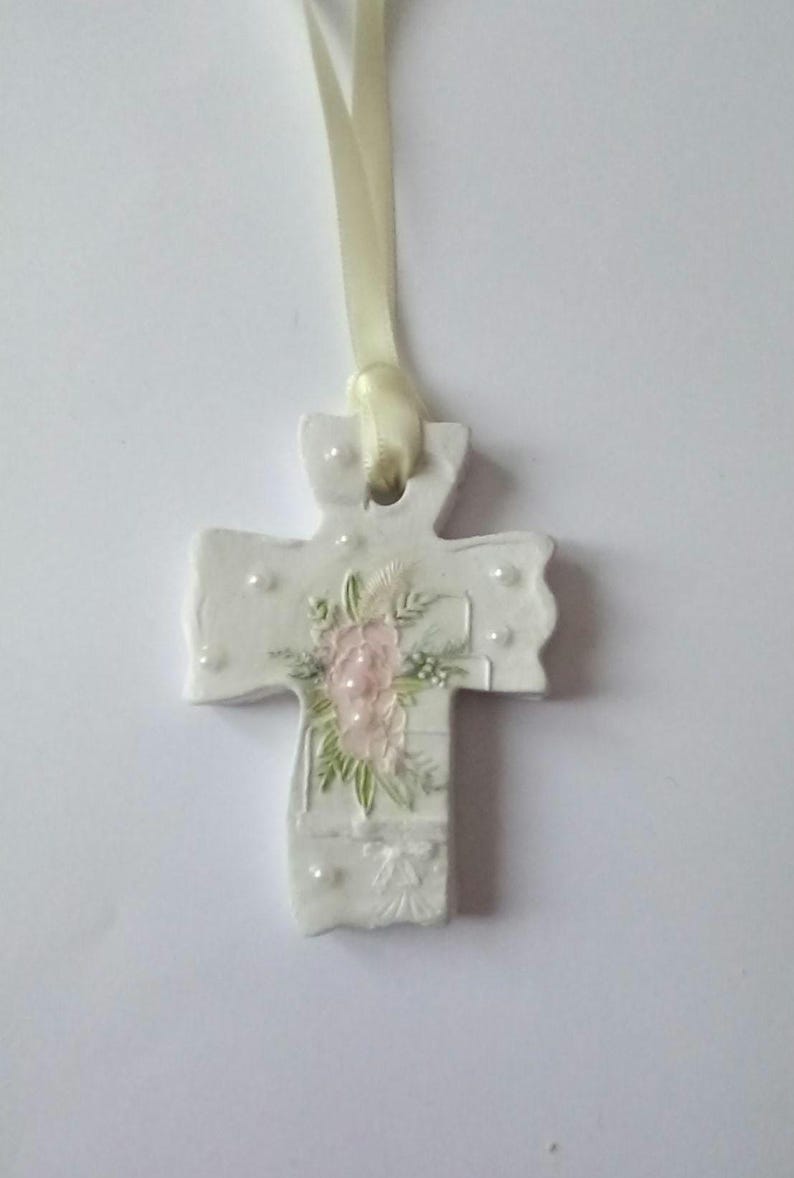 May include: A white ceramic cross pendant with a cream-colored ribbon. The cross is decorated with a pink floral design and small white pearl accents. The pendant is approximately 3 inches in length.