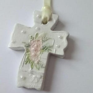 May include: White cross-shaped ornament with a floral design and pearl accents. The cross features a light pink flower arrangement with green leaves. A cream-colored ribbon is attached for hanging.