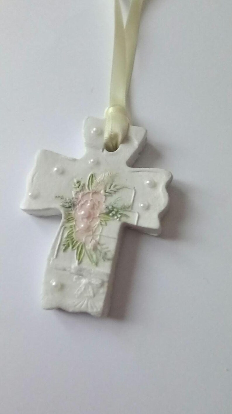 May include: White cross-shaped ornament with a floral design and pearl accents. The cross features a light pink flower arrangement with green leaves. A cream-colored ribbon is attached for hanging.