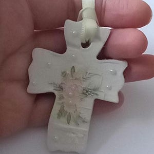 May include: A white ceramic cross ornament with a floral design in the center. The cross has a cream-colored ribbon for hanging. The floral design features pink and green hues. Small, clear embellishments are scattered across the surface.