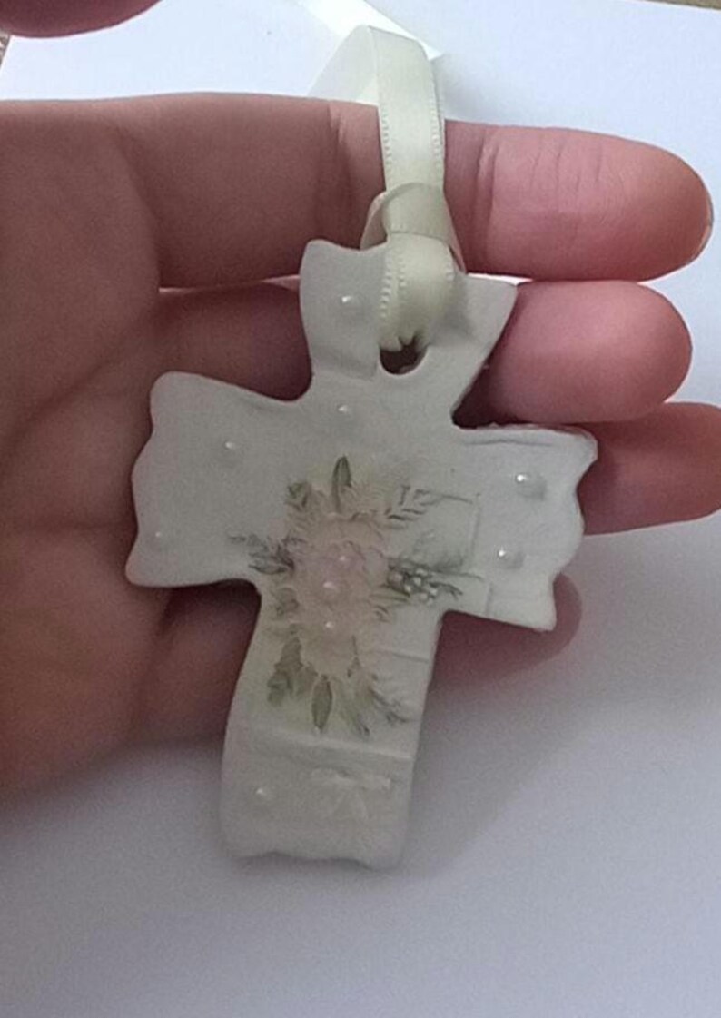 May include: A white ceramic cross ornament with a floral design in the center. The cross has a cream-colored ribbon for hanging. The floral design features pink and green hues. Small, clear embellishments are scattered across the surface.