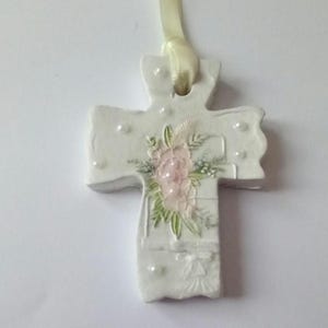 May include: A white ceramic cross pendant with a floral design in pink and green. The cross is adorned with small pearl-like accents and a cream-colored ribbon for hanging. A delicate religious ornament.