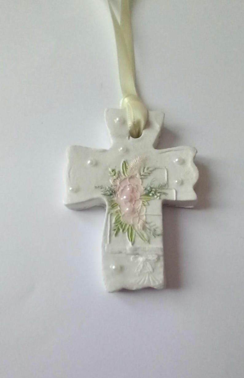 May include: A white ceramic cross pendant with a floral design in pink and green. The cross is adorned with small pearl-like accents and a cream-colored ribbon for hanging. A delicate religious ornament.