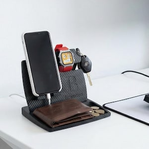 Könnte beinhalten: Ein schwarz-grauer Schreibtisch-Organizer mit Telefon, Uhr, Schlüsseln, Geldbörse und Münzen. Der Organizer hat einen Ladeanschluss für das Telefon. Ein Computermonitor, eine Tastatur und ein Mauspad befinden sich ebenfalls auf dem weißen Schreibtisch.