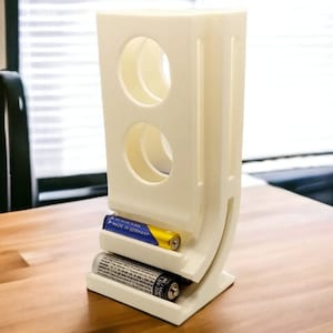 May include: A white 3D printed battery holder with two compartments for AA batteries. The holder has a curved base and a slot for holding batteries. Two AA batteries are in the holder, one on top of the other. The top battery has the text "MADE IN GERMANY" printed on it.