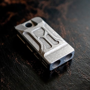 Könnte beinhalten: Ein kleines, rechteckiges, metallisch graues Werkzeug mit strukturierter Oberfläche. Das Werkzeug hat zwei kreisförmige Öffnungen an einem Ende und ein einzelnes Loch am anderen. Das Design umfasst geometrische Ausschnitte und Rillen.