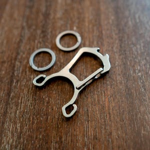 May include: A silver multi-tool with a bottle opener, key ring loops, and a carabiner-style clip. Two additional silver key rings are also present. The items are arranged on a dark wood surface.
