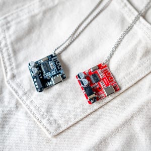May include: Two circuit board pendants on silver chains. One pendant is black, the other is red. The pendants are square with various electronic components. The necklaces rest on a white fabric surface.