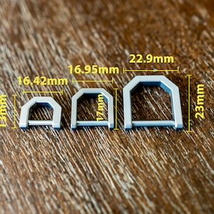 Op de afbeelding: Drie zilverkleurige metalen D-ringen van verschillende maten worden getoond op een houten oppervlak. De kleinste is 13 mm hoog en 16,42 mm breed. De middelste ring is 17 mm hoog en 16,95 mm breed. De grootste is 23 mm hoog en 22,9 mm breed.