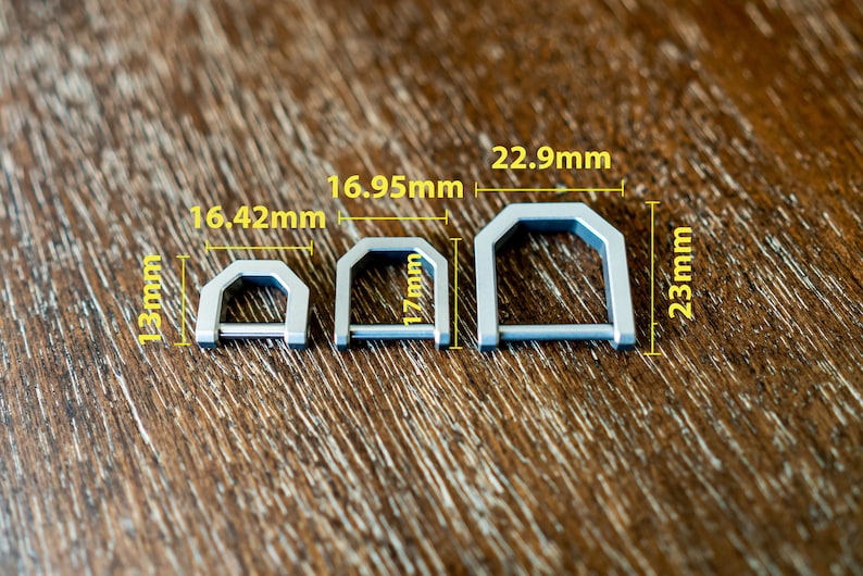 Op de afbeelding: Drie zilverkleurige metalen D-ringen van verschillende maten worden getoond op een houten oppervlak. De kleinste is 13 mm hoog en 16,42 mm breed. De middelste ring is 17 mm hoog en 16,95 mm breed. De grootste is 23 mm hoog en 22,9 mm breed.