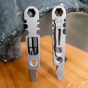 Könnte beinhalten: Zwei silberne Multi-Tools werden gezeigt. Das Werkzeug links hat einen Schlitz mit zwei Bits, einen Flaschenöffner und eine sechseckige Öffnung. Das Werkzeug rechts hat einen Taschenclip und einen Flaschenöffner. Beide Werkzeuge liegen auf einer Holzoberfläche.