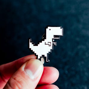 May include: A white and black pixelated dinosaur pin. The pin features a retro, 8-bit design of a Tyrannosaurus Rex. The dinosaur is set against a dark background. The pin is held by a person's fingers.