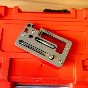 Puede incluir: Una herramienta multifunción rectangular de metal gris que presenta una ranura central con una punta de destornillador, un abrebotellas, un recorte hexagonal y un borde dentado. La herramienta está construida con varios tornillos pequeños.