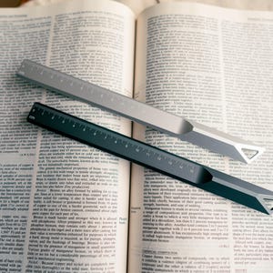 Könnte beinhalten: Zwei Metalllineale, eines silberfarben und eines dunkelgrau, liegen auf einem offenen Buch. Die Lineale haben einen dreieckigen Ausschnitt an einem Ende und sind mit Zentimetern markiert. Die Seiten des Buches sind mit kleinem Text gefüllt.