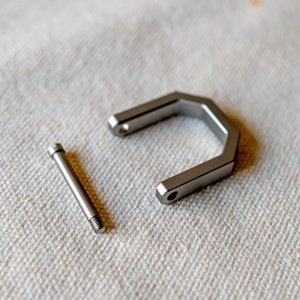 Op de afbeelding: Een gedemonteerde metalen componentenset, met een U-vormig stuk en een schroef. De stukken zijn zilverkleurig en lijken van roestvrij staal te zijn. De items liggen op een wit, getextureerd oppervlak.
