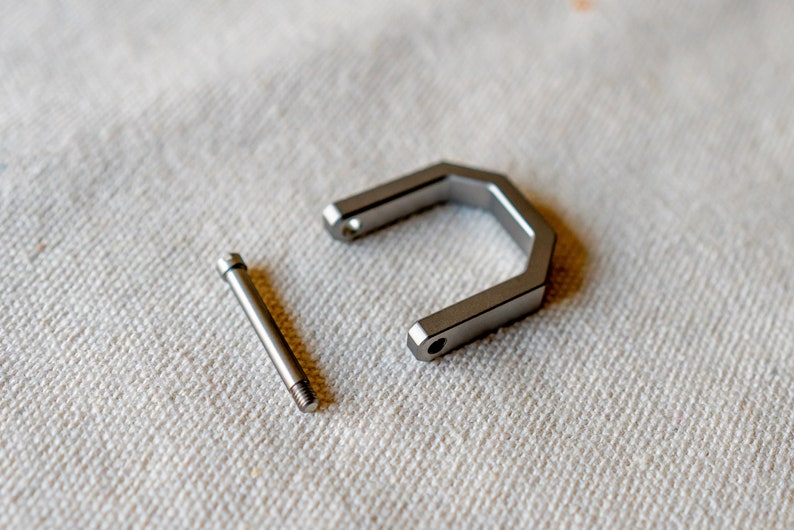 Op de afbeelding: Een gedemonteerde metalen componentenset, met een U-vormig stuk en een schroef. De stukken zijn zilverkleurig en lijken van roestvrij staal te zijn. De items liggen op een wit, getextureerd oppervlak.