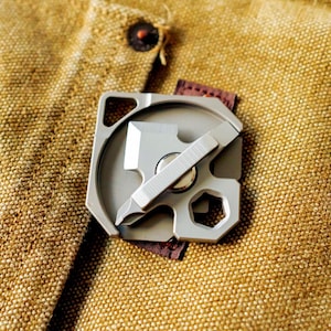 Puede incluir: Una multiherramienta plateada con cuchilla, destornillador y llave se muestra sobre una tela beige texturizada. La herramienta metálica compacta tiene un acento de cuero marrón. Está diseñada para diversas tareas.