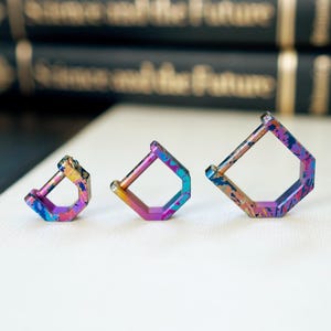 Puede incluir: Tres mosquetones cuadrados multicolores de diferentes tamaños. Los clips tienen un acabado iridiscente con tonos azules, morados y dorados. Están dispuestos sobre una superficie blanca, con libros borrosos en el fondo.