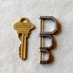 Op de afbeelding: Een messing Schlage-sleutel naast een zilverkleurige, geometrische sleutelhanger. De sleutelhanger is gemaakt van drie verbonden D-vormige elementen. Beide objecten liggen op een getextureerd, gebroken witte ondergrond.