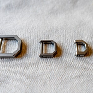 Op de afbeelding: Drie zilverkleurige D-ringgespen van verschillende maten liggen op een getextureerd, gebroken wit oppervlak. De gespen hebben een geometrisch ontwerp met een rechte staaf die twee zijden van een D-vormig frame verbindt.