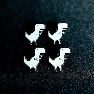 Könnte beinhalten: Vier pixelige, weiße Dinosaurier-Anstecknadeln auf einem dunklen, strukturierten Hintergrund. Jede Nadel zeigt ein vereinfachtes T-Rex-Design, das an Retro-Videospiele erinnert. Die Nadeln sind gleichmäßig angeordnet und bilden ein symmetrisches Muster.