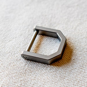 Op de afbeelding: Een zilverkleurige, D-vormige metalen gesp met een rechte staaf. De gesp heeft een geometrisch ontwerp met een zeshoekige vorm en een afgeronde staaf. Het item wordt getoond op een getextureerd, gebroken witte ondergrond.