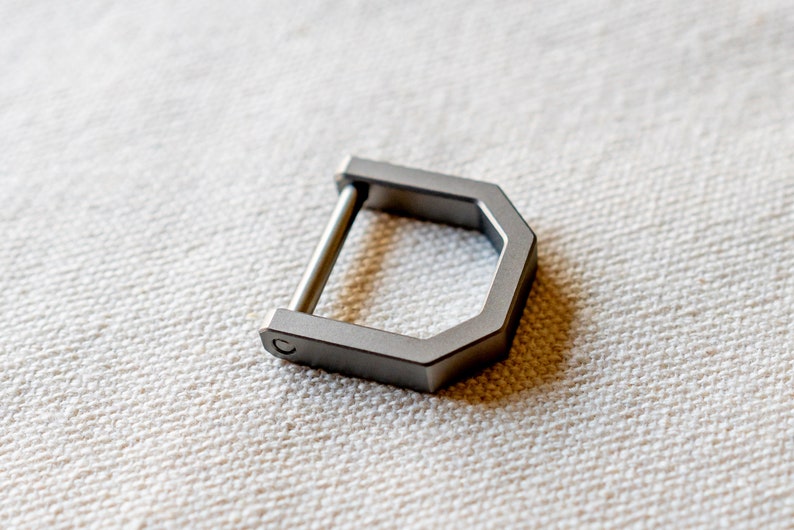 Op de afbeelding: Een zilverkleurige, D-vormige metalen gesp met een rechte staaf. De gesp heeft een geometrisch ontwerp met een zeshoekige vorm en een afgeronde staaf. Het item wordt getoond op een getextureerd, gebroken witte ondergrond.