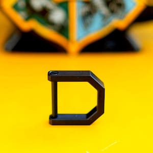 May include: A black metal D-ring carabiner with a rectangular shape and a straight bar. The carabiner is set against a bright yellow background. The carabiner is a hardware item.