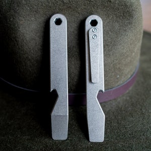 May include: Two silver metal multi-tools, one with a pocket clip, against a green hat. Each tool has a bottle opener and a flathead screwdriver. The tools have a hexagonal hole at the top.