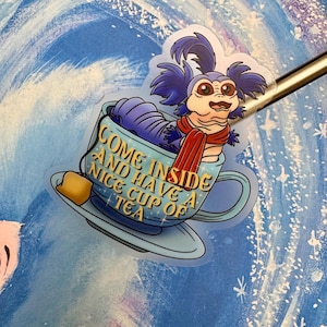 May include: A blue and purple cartoon worm wearing a red scarf sits in a blue teacup with a saucer. The text on the cup reads "Come inside and have a nice cup of tea."