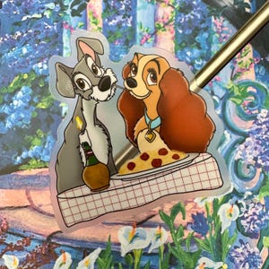 Op de afbeelding: Een sticker met Lady en de Tramp die een bord spaghetti delen. Lady is een bruine en witte hond met een rode halsband. Tramp is een grijze en witte hond met een bruine halsband. De sticker is op een roze en witte geruite tafelkleed.