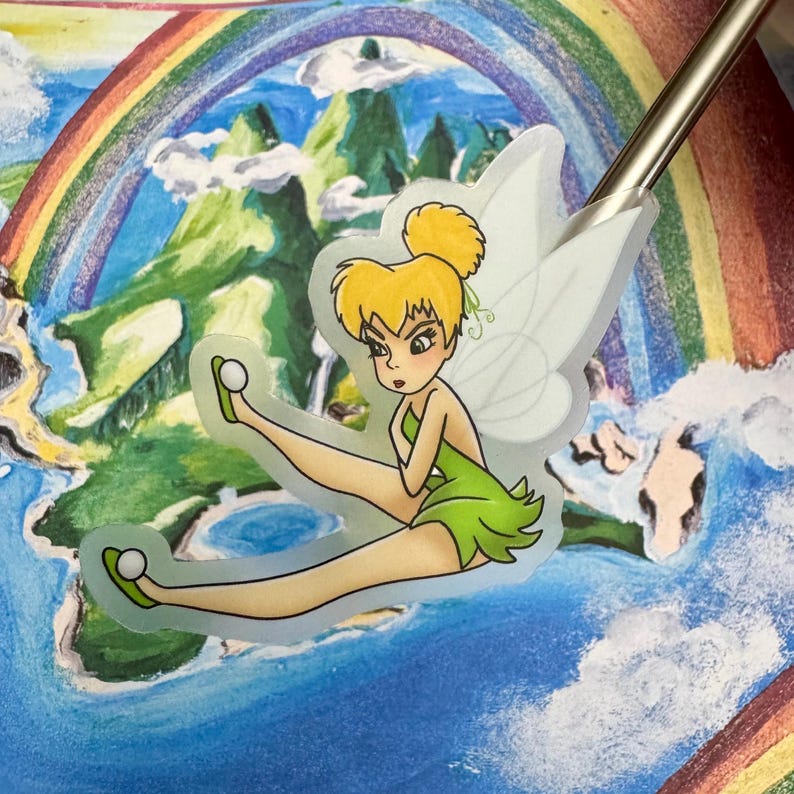 May include: A sticker featuring Tinker Bell, a fairy with blonde hair and green wings, flying over a rainbow and a green landscape.
