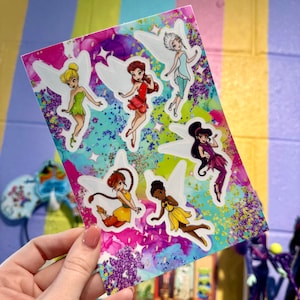 May include: A colourful sticker sheet featuring six fairies with wings. The fairies are wearing different coloured dresses and have different hairstyles. The background is a sparkly, glittery design.