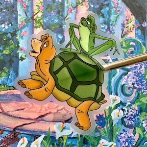 May include: A green frog sits on the back of a brown turtle with a green shell. The turtle is walking forward with its legs outstretched. The frog is looking to the left with its mouth open.